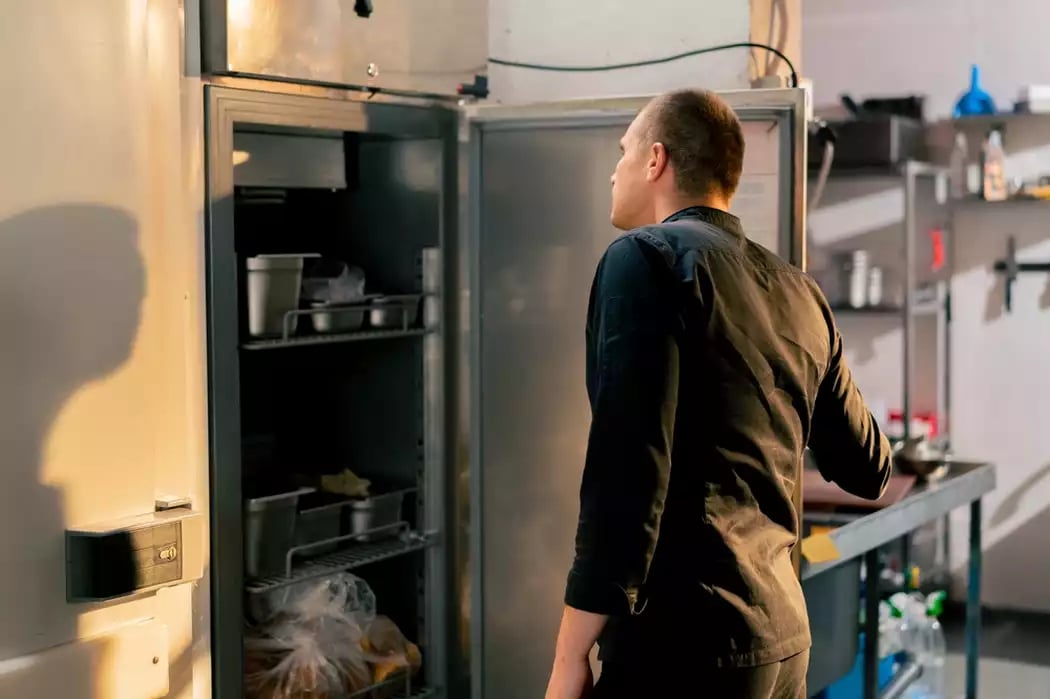 A chef in a uniform looking in a refrigerator and managing food waste in a restaurant.
