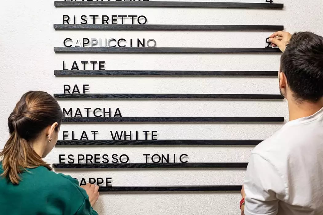 Two baristas adding restaurant menu pricing to a large letterboard menu on the wall.