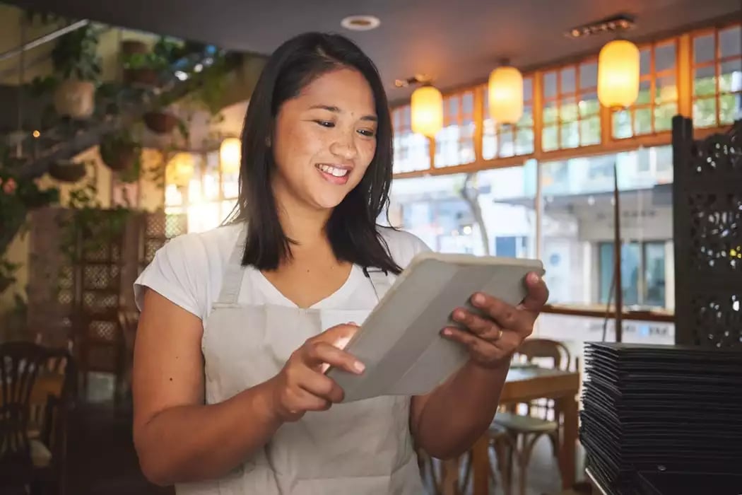 A young woman in an apron working on inventory on a tablet in a restaurant.